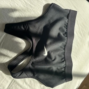 Svart sport-bh från Nike - Säljer en svart sport-bh från Nike med klassisk logga framtill. Modellen har brottarrygg och bred resår nedtill för extra stöd. Perfekt för träning och sportiga aktiviteter.