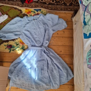 Ljusblå kortklänning med volang och bälte - Superfin ljusblå klänning med kort ärm och volangdetaljer. Klänningen har ett knytbart bälte i midjan och är tillverkad i ett luftigt material som passar perfekt till varma dagar. Söt och trendig modell med rund halsringning.