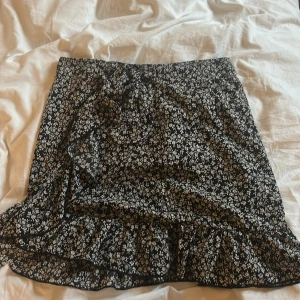 Svart blommig omlottkjol med volang - Säljer en svart omlottkjol med vitt blommönster och volangkant. Kjolen har knyt i midjan och är perfekt för dig som gillar en feminin och trendig stil.