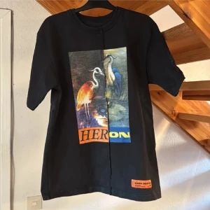 Svart t-shirt med fågelmotiv från Heron Preston - Cool svart t-shirt från Heron Preston med stort färgglatt tryck av hägrar på framsidan och orange logotypdetalj nertill. Klassisk passform och rund halsringning. Perfekt statement-plagg för dig som gillar streetwear. Storleken är L oversized.