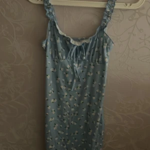 Blommig blå klänning med volangdetaljer - Söt blå klänning med små vita blommor och gröna blad. Klänningen har volang vid axelbanden och en knytning framtill vid bysten. Perfekt för varma dagar och har en romantisk känsla.