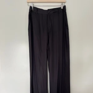 Svarta byxor, Rabalder, M - Eleganta, svarta byxor med vida ben från Rabalder. Byxorna har en klassisk design med dragkedja och knappstängning framtill, samt praktiska fickor på sidorna. Perfekt för en stilren och bekväm look. Små diskreta fickor bak. Öglor för skärp finns också. Byxorna är i nyskick. Sidenliknande material. 100% viskos.   Mått: Midja ca 40 cm Stuss ca 37 cm Längd yttre ca 103 cm Längd inre ca 71 cm