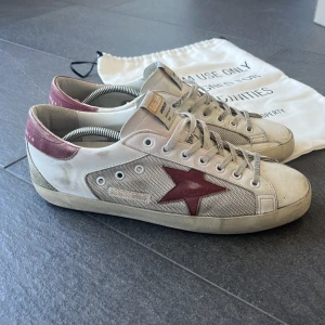 Golden Goose Super-Star   - Säljer ett par vita Golden Goose sneakers med röd stjärndetalj på sidan och vinröda hälpartier. Bra skick och riktigt snygga inför sommaren! Skopåse och skokartong ingår då de är köpta från miinto.se 