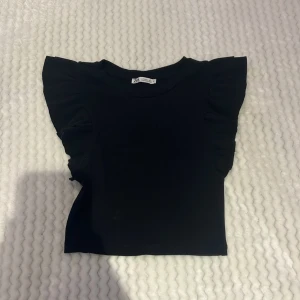 Svart croppad topp med volang från Zara - Säljer en svart croppad topp från Zara med volangdetaljer vid axlarna. Toppen är ribbad och har en tight passform.