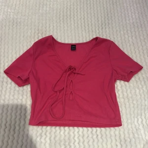 Rosa ribbad croppad topp från SHEIN - Säljer en rosa croppad topp från SHEIN med ribbad struktur och v-ringning. Toppen har korta ärmar och knytning framtill som en snygg detalj. Perfekt för vår och sommar!