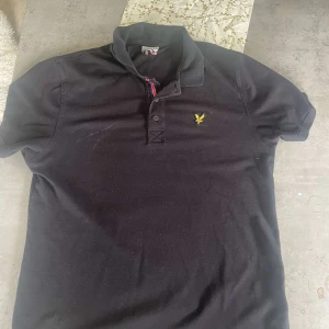Svart piké t-shirt från Lyle & Scott - Klassisk svart piké t-shirt från Lyle & Scott med korta ärmar och krage. Bröstet pryds av den ikoniska gula loggan. Tillverkad i bomull för en skön känsla. Perfekt för en stilren och avslappnad look.