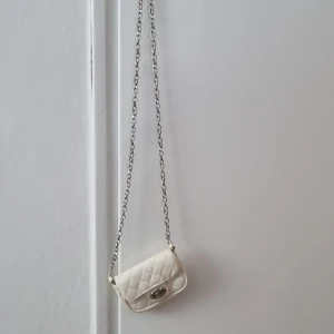 Vit quiltad mini axelväska med kedja - Säljer en liten vit quiltad axelväska från att. Väskan har silverfärgad kedja och ett snyggt vridlås framtill. Perfekt för det viktigaste och ger en trendig touch till din outfit. Student väska