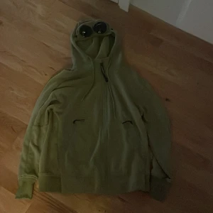 Grön hoodie med goggles från C.P. Company - Säljer en grön hoodie från C.P. Company med deras ikoniska goggles i huvan. Tröjan har dragsko i huvan och två fickor framtill. Perfekt för dig som vill sticka ut med en unik streetwear-look.