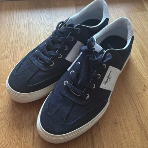 Mörkblå och vita sneakers från Pepe Jeans - Säljer ett par stilrena mörkblå sneakers från Pepe Jeans med vita detaljer och vit sula. Skorna har snörning, rund tå. Perfekta för dig som gillar en klassisk och sportig look. Använda endast en gång!