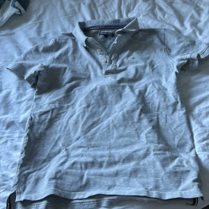 Ljusgrå piké t-shirt från Tommy Hilfiger - Storlek 164 skulle säga att det är som XS                                                                                   Säljer en ljusgrå piké t-shirt från Tommy Hilfiger med klassisk krage och knappar framtill. Liten broderad logga på bröstet och korta ärmar. Perfekt för en varm dag i solen.