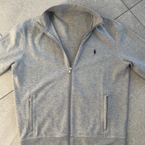 Grå ziptröja från Polo Ralph Lauren - Säljer en stilren grå ziptröja från Polo Ralph Lauren med klassisk broderad logga på bröstet. Tröjan har hög krage, dragkedja framtill och två fickor. Perfekt för lager-på-lager och enkel att matcha.