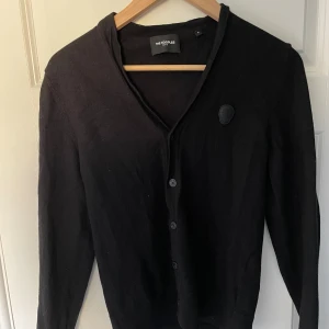 The Kooples Paris Cardigan - Säljer en svart Cardigan från The Kooples med knäppning framtill och v-ringad hals. Tröjan har långa ärmar och snygga detaljer med svarta armbågslappar. Tröjan är lite urtvättad så har krympt lite i tvätten.