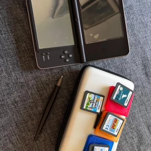 Nintendo DSi XL med spel och fodral - Nintendo DSi XL i svart med tillhörande stylus, flera spelkassetter och ett vitt fodral. Den fungerar som den ska men laddare saknas!   