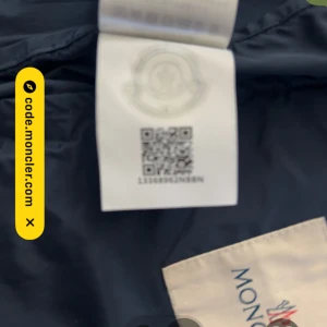 Mörkblå vindjacka från Moncler - Säljer en mörkblå vindjacka från Moncler med huva och dragkedja framtill. Jackan har två fickor med tryckknappar och en vit Moncler-logga på bröstet. Klassisk design med röd och vit rand på huvan.