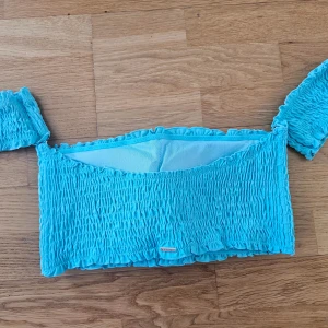 Turkos smockad offshoulder topp - Säljer en turkos smockad topp/ bikiniöverdel med offshoulder-design och korta ärmar. Toppen har en croppad passform och är  stretchig. Passar till M upptill XL.  Avtagbara band! Aldrig använd! Helt ny, men etiketten har jag tagit bort 