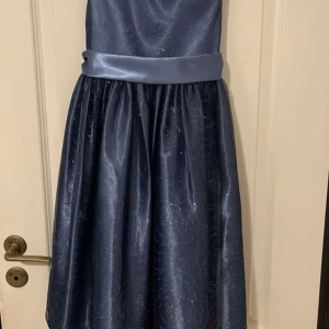 Marinblå långklänning med rosett från LOVE byspecialoccasions - Elegant marinblå långklänning i glansig polyester med bred satinmidja och stor rosett bak. Klänningen har ärmlös design och subtilt mönster i tyget som ger en lyxig känsla. Perfekt för festliga tillfällen.