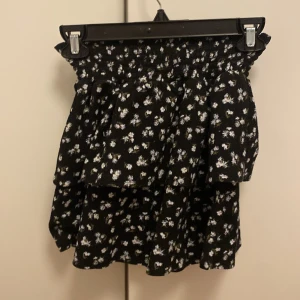 Svart blommig kjol från Kappahl - Supersöt svart kjol från Kappahl med vitt blommönster och volangdetaljer. Kjolen har en smockad midja som ger en bekväm passform och en luftig känsla. Perfekt för dig som gillar en feminin och trendig stil.