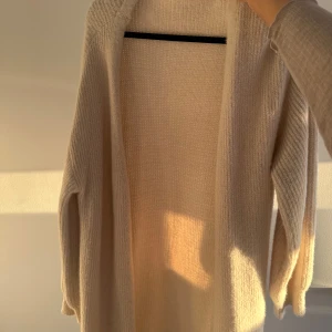 Beige stickad kofta - Storlek S. Säljer en lång, öppen beige stickad kofta med långa ärmar. Koftan är mjuk och har en avslappnad passform, perfekt att slänga på sig när det är lite kyligt ute.