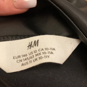 Svart skinnkjol från H&M - En svart, enkel kjol från H&M i klassisk rak modell. Kjolen har en normal passform och är knälång, vilket gör den lätt att matcha med olika toppar. Perfekt för dig som gillar stilrena plagg.