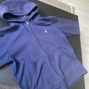 Marinblå hoodie från Polo Ralph Lauren - Säljer en marinblå hoodie från Polo Ralph Lauren med dragkedja och huva. Tröjan har två fickor framtill och den klassiska broderade loggan på bröstet. Perfekt för en avslappnad stil. Pris går att diskuteras. Kom privat för mer info