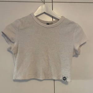 Vit croppad t-shirt från Divided - Säljer en vit croppad t-shirt från Divided med korta ärmar och rund halsringning. Materialet är frottéliknande och har en liten Cozy Wave Surf Club-lapp nertill. 