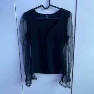 Svart topp med genomskinliga ärmar. - Säljer en svart topp från SHEIN med långa genomskinliga meshärmar. Toppen har en v-ringad hals och en jättefin passform. Perfekt för dig som vill ha något stilrent med en twist, tänk denna festliga tröja med smycken kommer verkligen att poppa till! (Pris kan diskuteras)