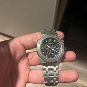 Silverfärgad kronografklocka från Regal - Stilren herrklocka från Regal i silverfärgat rostfritt stål med länkarmband och svart urtavla. Klockan har kronograffunktion och tydliga indexmarkeringar. Snygg design med åttakantig boett och detaljerade knappar på sidan.