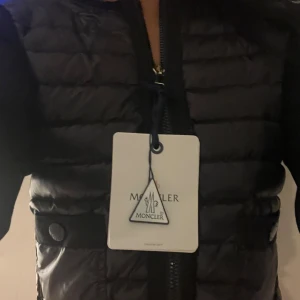 Svart cardigan från Moncler - Säljer en svart dunväst från Moncler med quiltad design och dragkedja framtill. Västen har Moncler-logga på bröstet och ribbade detaljer vid axlar och sidor. Perfekt för lager-på-lager och snygg till många outfits.