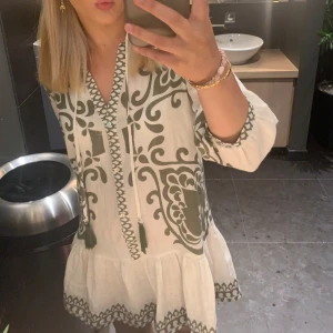Zara klänning med mönster - Säljer en luftig vit klänning med grönt grafiskt mönster och v-ringning. Klänningen har trekvartsärm och tofsdetaljer vid halsen. Perfekt för varma dagar och har en avslappnad passform.💕💕