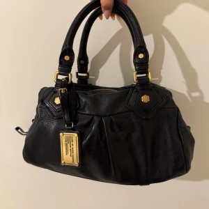 Svart handväska från Marc by Marc Jacobs - Säljer en svart handväska från Marc by Marc Jacobs med guldfärgade metalldetaljer och dragkedja. Väskan har två rejäla handtag och en dekorativ metallbricka framtill. Tillverkad i skinn med mjuk, något rynkad form. Använd o kommer med förlängning. Skador finns i bilderna❤️