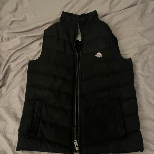 Svart dunväst från Moncler - Säljer en svart dunväst från Moncler med hög krage och dragkedja framtill. Västen har två fickor med dragkedja och Moncler-logga på bröstet. Storlek M. Dragkedja fungerar inte och stygnen håller på o lossna till den lilla animerade rutan på insidan