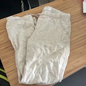 Beiga linnebyxor från Lindex - Snygga och luftiga beiga linnebyxor från Lindex med resår i midjan och dragsko. Perfekta för varma dagar och har en avslappnad passform med raka ben. Byxorna har sidfickor och är tillverkade i ett tunt, svalt material. STORLEK: EUR -UK 11-12Y STORLEKEN NEDANFÖR STÄMMER TROLIGEN EJ!