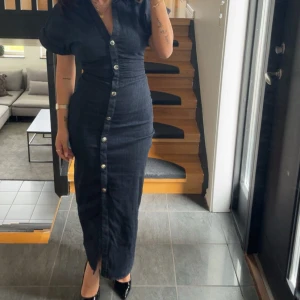 Mörkblå jeansklänning med knappar - Snygg lång mörkblå jeansklänning med korta ärmar och markerad midja. Klänningen har knappar hela vägen framtill och en liten slits nertill. Perfekt för dig som gillar en klassisk men trendig look.