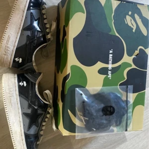 Svarta BAPE sneakers med påse - Säljer ett par svarta BAPE sneakers med glansig finish och vit sula. Skorna har den klassiska stjärndetaljen på sidan och BAPE-loggan på hälen. Levereras med originalkartong i camomönster och extra skosnören.