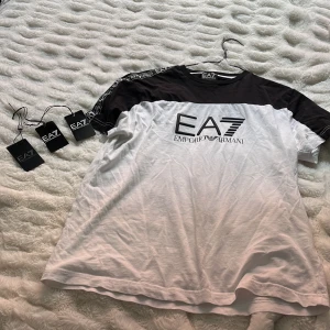 Svartvit t-shirt från EA7 Emporio Armani - Säljer en snygg svartvit t-shirt från EA7 Emporio Armani med stor logga framtill och svarta axelband med EA7-tryck. T-shirten har korta ärmar och är gjord i mjuk bomull. Perfekt för en trendig och sportig look. Köpt på urban modern hallarna Halmstad för 1200kr