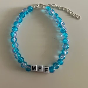 Blått pärlarmband med MFF-bokstäver - Snyggt armband med blå och genomskinliga pärlor samt vita kubpärlor med svarta bokstäver som bildar 'MFF'. Justerbar kedja i silverfärg. Perfekt accessoar för dig som vill visa ditt stöd för MFF Mått utan länk: 17 cm Mått med länk: 22 cm