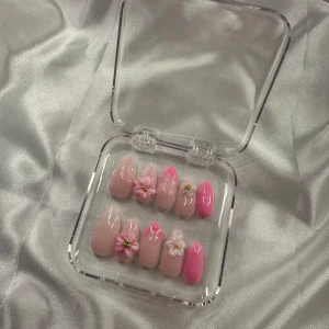 Press-on naglar med blommor och rosa detaljer - Säljer ett set med press-on naglar i olika nyanser av nude och rosa. Några naglar har 3D-blommor och små pärlor som dekoration. Naglarna är långa och har en spetsig form, levereras i en genomskinlig ask.