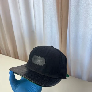 Doses keps - Snygg svart keps från Doses med krokodilpräglad skärm och justerbar baksida. Skärmen är svart på ovansidan och grön undertill. Framsidan har en svart patch med broderad logga.