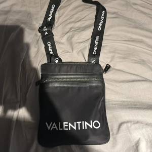 Svart axelväska från Valentino - Säljer en svart axelväska från Valentino med justerbart axelband där märkesnamnet står tryckt. Väskan har två dragkedjefack och en stilren design med logga framtill. Perfekt för att bära det viktigaste i vardagen.