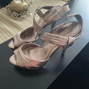 Champagnefärgade klackskor med öppen tå - Säljer ett par eleganta champagnefärgade klackskor från Menbur. Skorna har öppen tå, hög klack och breda remmar som korsar över foten och runt vristen. Perfekta för dig som vill ha en stilren och festlig look.