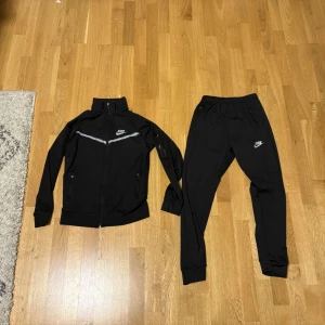 Svart träningsset från Nike - Säljer ett svart träningsset från Nike med matchande jacka och byxor. Jackan har dragkedja, ståkrage och en vit detalj över bröstet samt Nike-logga. Byxorna har resår i midjan och Nike-logga på låret. Använd 1-2 gånger! Pris kan diskuteras!😉