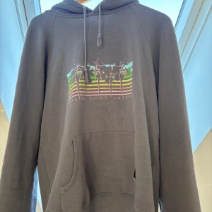Grå hoodie från Saint Laurent med palmmotiv - Grå limited edition hoodie från Saint Laurent med färgglatt palmmotiv och texten 'Malibu Saint Laurent' på bröstet. Tröjan har huva med dragsko och en stor magficka framtill. Perfekt för en avslappnad och cool stil.