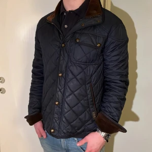 Svart quiltad jacka med bruna detaljer - Snygg svart quiltad jacka med bruna manchesterdetaljer på krage och ärmslut. Jackan har dragkedja och knappar framtill samt fickor med lock och knapp. Perfekt för dig som vill ha en stilren och klassisk look.