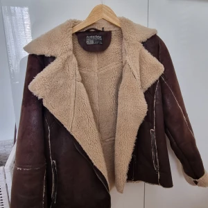 Brun pälsfodrad jacka från Saints & Mortals - Säljer en brun jacka från Saints & Mortals i storlek L. Jackan har ett mjukt, ljust teddyfoder och stor krage. Yttermaterialet är mockaliknande och jackan har dragkedjor vid fickorna. Perfekt för kyliga dagar. Läderjacka