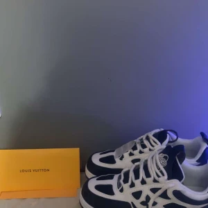 Louis Vuitton sneakers i marin blå och vitt - Säljer ett par Louis Vuitton sneakers i marin blå och vitt med klassiska LV-detaljer och logga på hälen. Skorna har snörning, rund tå och platt sula. Perfekta för dig som vill ha en lyxig och trendig look.