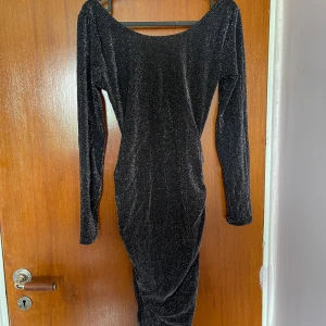 Svart glittrig klänning med öppen rygg - Säljer en svart långklänning med glittrigt tyg och lång ärm. Klänningen har en snygg öppen rygg med kedjedetalj och draperad kjol. Perfekt för dig som vill sticka ut på festen!