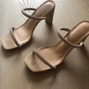 Beige klackskor med glittriga remmar  - Säljer ett par snygga beige klackskor  med fyrkantig tå och två smala glittriga remmar. Skorna har en stabil blockklack och öppen tå, perfekta för att lyfta din outfit. Passar storlek 38-40