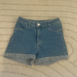 Blå jeansshorts från H&M - Snygga blå jeansshorts från H&M med klassisk femficksdesign och uppvikta benslut. Perfekta för varma dagar och enkla att matcha med olika toppar. Stängs med knapp och dragkedja framtill.