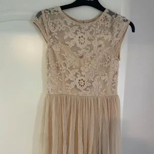 Champagnefärgad spetsklänning med volangkjol - Romantisk champagnefärgad klänning med blommig spets upptill och volangig kjol i tunt tyg. Klänningen har korta ärmar, rund halsringning och öppen rygg. Perfekt för sommarens speciella tillfällen.
