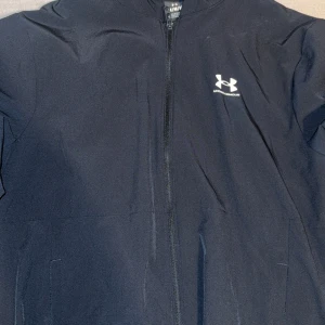 Svart vindjacka från Under Armour - Säljer en svart vindjacka från Under Armour med huva och dragkedja framtill. Jackan har lång ärm och diskret logga på bröstet. Perfekt för träning eller när du vill ha en lätt jacka med sportig känsla.
